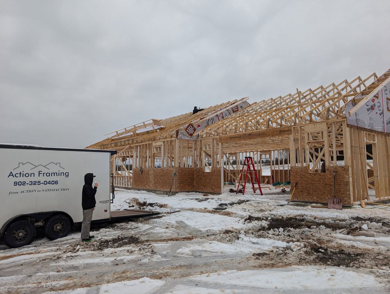 Long bungalow with roof rafters framed, snow on the ground and the Action Framing trailer alongside.