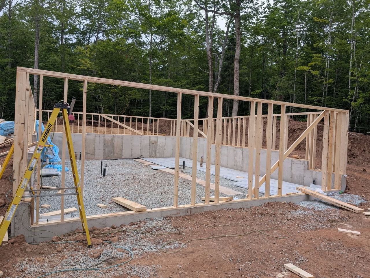 Exterior wall framing standing on a poured concrete foundation on a wooded lot.